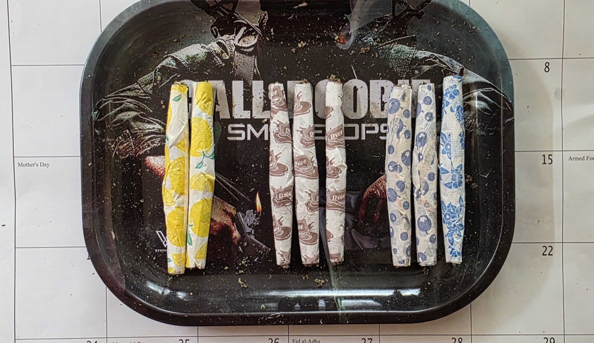 Three pairs of pre-rolled joints on a black rolling tray, sitting on a wall calendar showing Memorial Day weekend: yellow lemon-print papers on the left, brown chocolate-print Dvoe papers in the middle, blue blueberry-print papers on the right.